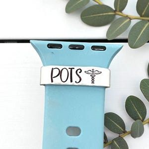 May include: A light blue watch band with a silver accent piece. The accent piece has the word "POTS" and a medical symbol. The watch band is on a white wooden surface with green leaves.