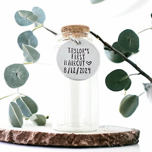 May include: A clear glass bottle with a cork top and a silver tag that reads "Taylor's First Haircut 8/12/2024".