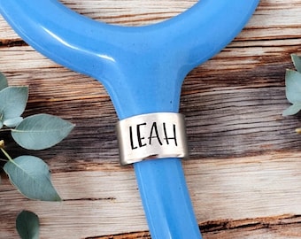 Könnte beinhalten: Ein blaues Stethoskop mit einem silbernen Namensschild, auf dem "LEAH" steht, auf einer Holzoberfläche.