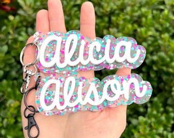May include: Two clear acrylic keychains with the names "Alicia" and "Alison" written in white script. The keychains are filled with pink, blue, and white glitter.