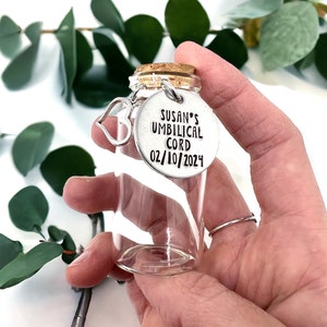 May include: A clear glass jar with a cork lid and a silver charm attached. The charm is a round disc with the text "SUSAN'S UMBILICAL CORD 02/10/2024".