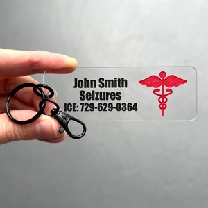 May include: A clear plastic keychain with a black and white text label that reads "John Smith Seizures ICE: 729-629-0364". A red medical symbol is printed on the right side of the keychain.
