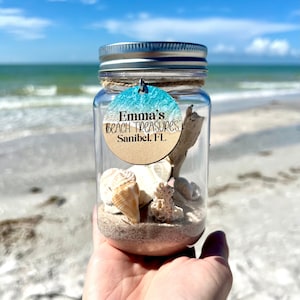 May include: A clear glass jar with a silver lid filled with sand, seashells, and a small piece of driftwood. A round wooden tag with a blue and white beach scene is attached to the jar with twine. The tag reads "Emma's Beach Treasures Sanibel, FL."