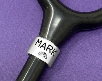 May include: A black stethoscope with a silver charm that says "MARK" with a rainbow underneath. The charm is a personalized stethoscope charm.