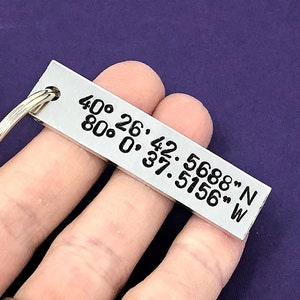 May include: Silver keychain with engraved coordinates, "40° 26'42.5688" N 80° 0'37.5156" W", on a purple background. The text "Personalized Coordinates Keychain" is visible above the keychain.