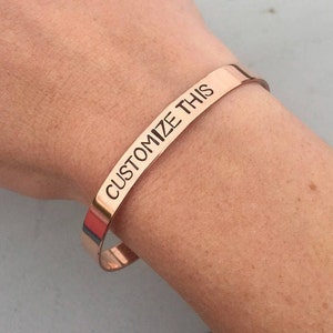 May include: A rose gold bangle bracelet with the words "CUSTOMIZE THIS" engraved on it.