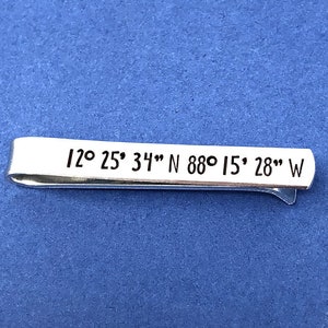 May include: Silver tie clip with the text "12° 25' 34" N 88° 15' 28" W" engraved on it. The tie clip is labeled "Gps Coordinates Tie Clip".