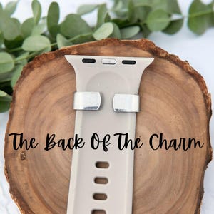 May include: A light beige Apple Watch band with silver-colored metal clasps. The band is displayed on a wooden surface with green foliage in the background. The text "The Back Of The Charm" is printed on the image.