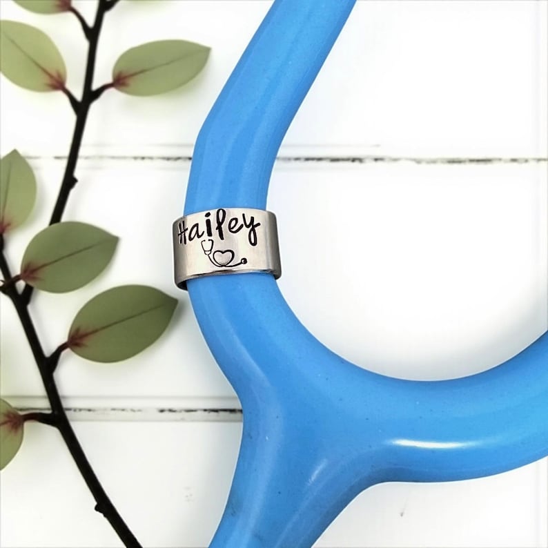 May include: A silver stethoscope charm with the name "Hailey" and a stethoscope graphic. The charm is attached to a blue stethoscope. Green leaves and a black branch are in the background.