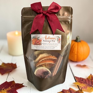 May include: A brown stand-up pouch with a dark red ribbon bow at the top. The label reads Autumn Simmer Pot and lists ingredients including dried orange, apple, cranberries, cinnamon stick, whole cloves, fig, and star anise.