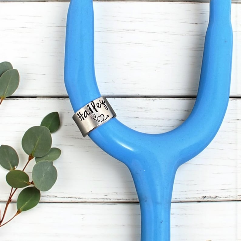 May include: A silver stethoscope charm with the name "Hailey" in cursive script and a small heart and stethoscope symbol. The charm is on a blue stethoscope against a white wooden background.