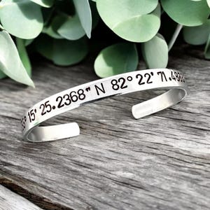 May include: A silver cuff bracelet with engraved coordinates. The bracelet is open-ended and sits on a wooden surface, with green foliage in the background. The engraved text includes numbers and symbols.