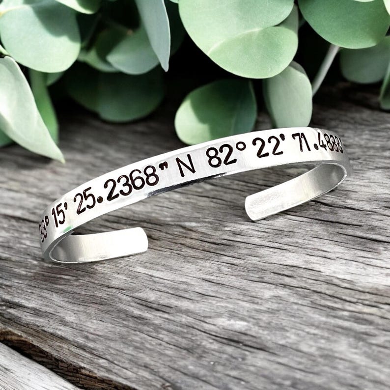 May include: A silver cuff bracelet with engraved coordinates. The bracelet is open-ended and sits on a wooden surface, with green foliage in the background. The engraved text includes numbers and symbols.