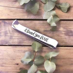 May include: Silver tie bar with the inscription "I loved you first" on a wooden surface with green leaves.