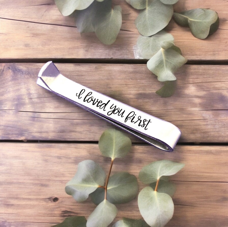 May include: Silver tie bar with the inscription "I loved you first" on a wooden surface with green leaves.
