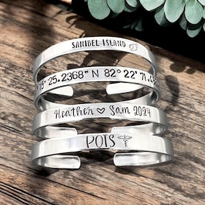 May include: Four silver-colored, open-ended bracelets. Each bracelet has engraved text: "SANIBEL ISLAND", coordinates, "Heather ♡ Sam 2024", and "POTS" with a medical symbol. The bracelets are displayed on a wooden surface.