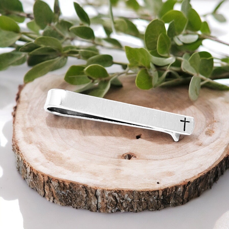 May include: Silver tie clip with a cross design. The tie clip is on a wooden surface.