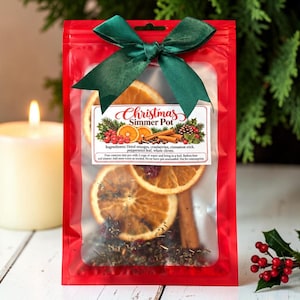 May include: A red plastic pouch containing dried orange slices, cranberries, and cinnamon sticks, tied with a dark green satin ribbon. A white label on the front reads Christmas Simmer Pot with instructions and a list of ingredients including dried oranges, cranberries, cinnamon stick, peppermint leaf, and whole cloves.