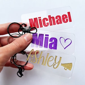 May include: Three clear acrylic keychains with names printed on them. The top keychain has the name "Michael" printed in red. The middle keychain has the name "Mia" printed in purple with a purple heart. The bottom keychain has the name "Ashley" printed in gold with a gold megaphone.