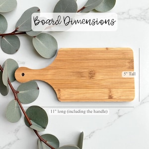 K&ouml;nnte beinhalten: Rechteckiges Schneidebrett aus Bambus mit Griff. Das Brett ist 12,7 cm hoch und 28 cm lang, einschlie&szlig;lich des Griffs. Der Text "Board Dimensions" steht oben. Eukalyptusbl&auml;tter im Hintergrund.
