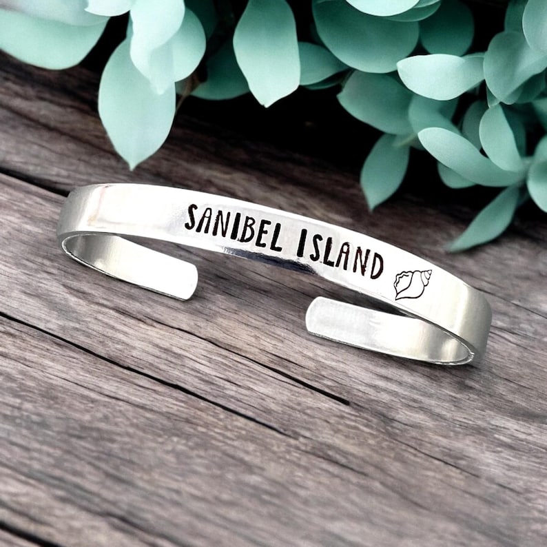 May include: A silver cuff bracelet with the words "SANIBEL ISLAND" in black lettering and a seashell graphic. The bracelet is open-ended and sits on a wooden surface with green foliage in the background.