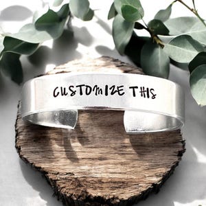May include: Silver metal cuff bracelet with the text "CUSTOMIZE THIS" engraved on the front.