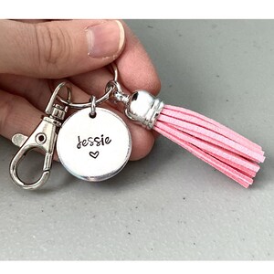 May include: A silver keychain with a pink tassel and a round charm that says "Jessie" with a heart. The keychain is a personalised gift.