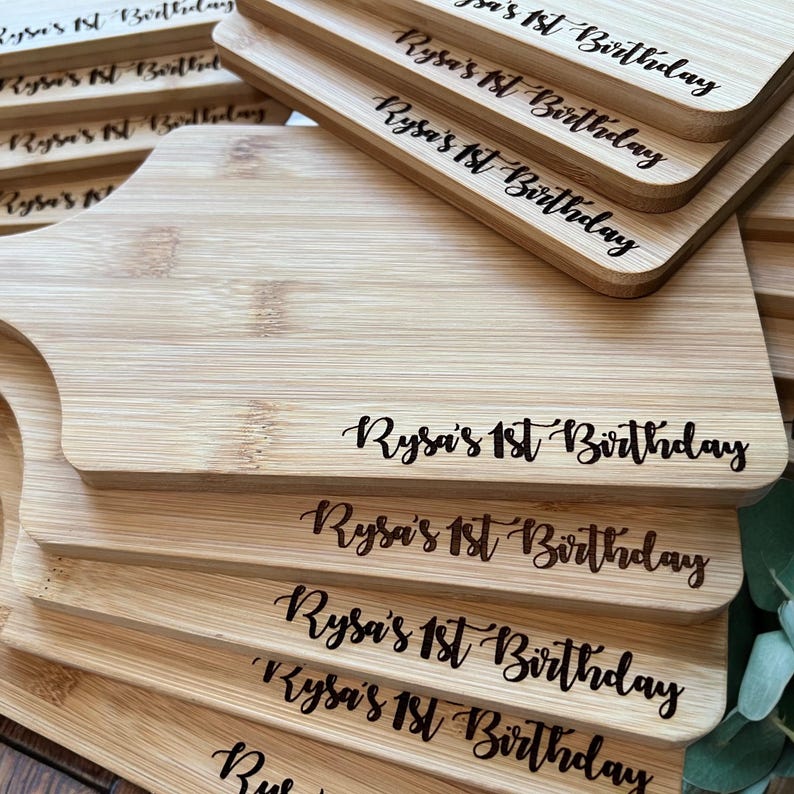 K&ouml;nnte beinhalten: Ein Stapel hellfarbiger Bambus-Schneidebretter. Jedes Brett hat eine abgerundete Kante und einen Griff. Die Worte "Rysa's 1st Birthday" sind in schwarzer Schrift auf jedes Brett ge&auml;tzt, passend f&uuml;r eine Feier.
