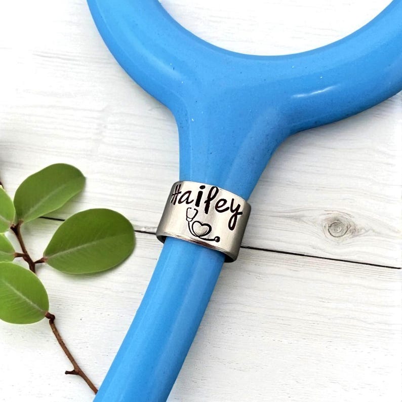 May include: A silver stethoscope ring with the name "Hailey" in a handwritten font and a small stethoscope graphic. The ring is wrapped around the blue handle of a stethoscope. Green leaves are visible in the background.