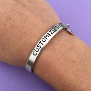 K&ouml;nnte beinhalten: Ein silbernes Armband mit der Gravur "CUSTOMIZE THIS". Das Armband hat einen kleinen Anh&auml;nger.