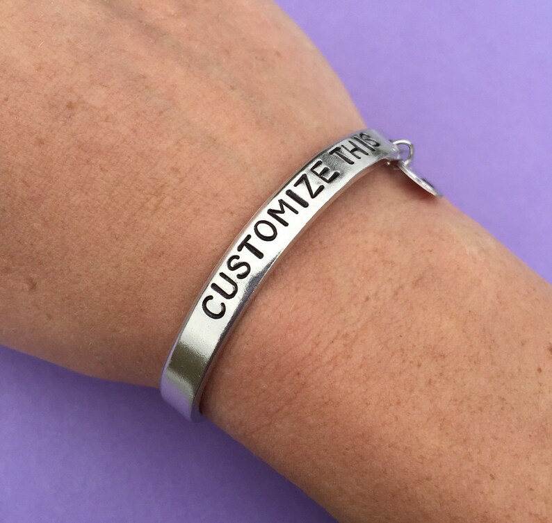 K&ouml;nnte beinhalten: Ein silbernes Armband mit der Gravur "CUSTOMIZE THIS". Das Armband hat einen kleinen Anh&auml;nger.