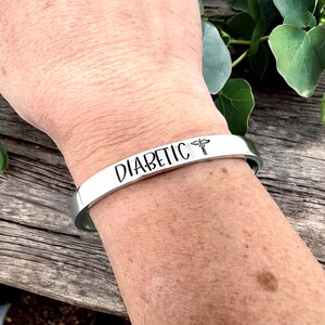 May include: A silver-colored cuff bracelet with the word "DIABETIC" in black lettering. A medical symbol is next to the word. The bracelet is worn on a wrist.
