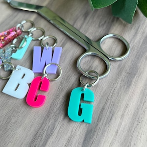 May include: A set of colorful letter charms with silver key rings. The letters are B, C, G, W, and a pink and white striped charm. The charms are on a wooden surface.