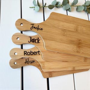 K&ouml;nnte beinhalten: Vier personalisierte Bambus-Schneidebretter mit Griffen. Jedes Brett ist mit einem Namen graviert: Jackie, Jack, Robert und Alison. Die Bretter sind hellbraun mit nat&uuml;rlicher Holzmaserung und einem Loch im Griff zum Aufh&auml;ngen.