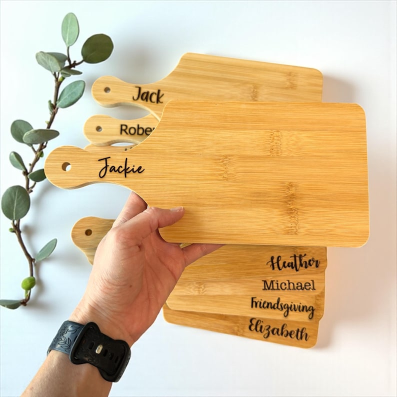K&ouml;nnte beinhalten: Mehrere personalisierte Schneidebretter aus Bambus. Jedes Brett hat einen Griff und ist mit Namen wie "Jack", "Robert" und "Jackie" graviert. Weitere Bretter tragen die Inschriften "Heather", "Michael", "Friendsgiving" und "Elizabeth".