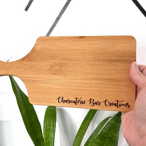 K&ouml;nnte beinhalten: Ein rechteckiges Schneidebrett aus Bambus mit Griff und kleinem Loch. Der Text "Charcuterie Bar Creations" ist in das Holz ge&auml;tzt. Das Brett wird von einer Person gehalten.