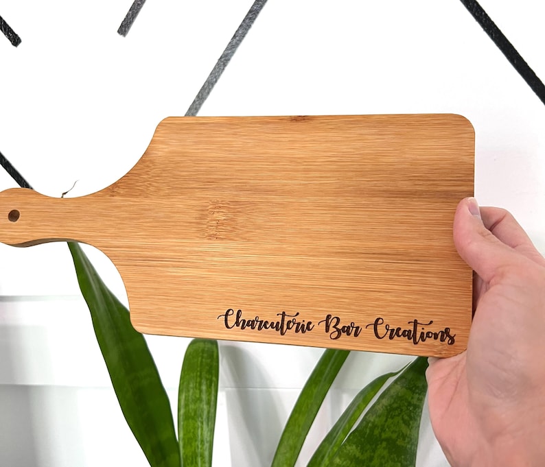 K&ouml;nnte beinhalten: Ein rechteckiges Schneidebrett aus Bambus mit Griff und kleinem Loch. Der Text "Charcuterie Bar Creations" ist in das Holz ge&auml;tzt. Das Brett wird von einer Person gehalten.