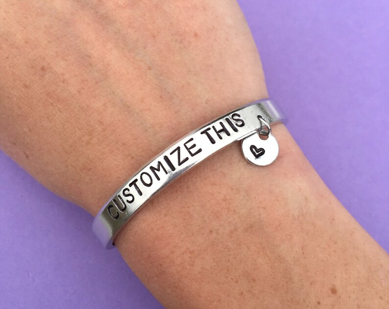 K&ouml;nnte beinhalten: Silberarmband mit der Gravur "CUSTOMIZE THIS". Ein kleiner silberner Anh&auml;nger mit einem Herzmotiv h&auml;ngt am Armband.