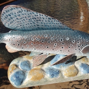 May include: A hand-carved wooden sculpture of a trout fish with brown and silver scales. The fish is perched on a base of painted rocks.