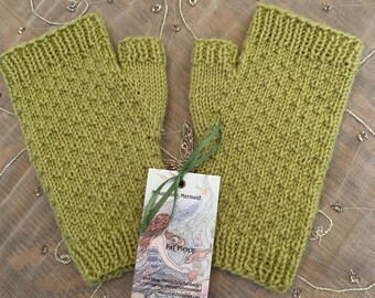 May include: A pair of olive green knitted fingerless gloves. The gloves are made of a textured knit fabric and have a tag attached with a mermaid illustration and the name "Pat Pierce". The gloves are displayed on a decorative surface.
