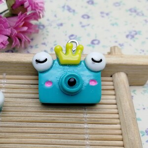 May include: Two turquoise blue camera charms with white eyes, pink cheeks, and a yellow crown. The charms have a silver loop for attaching to a necklace or bracelet.