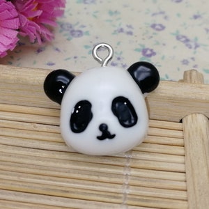 May include: A white ceramic panda charm with black eyes, nose, and ears. The charm has a silver loop for attaching to a necklace or bracelet.