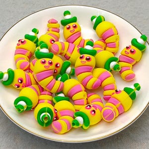 May include: A collection of colorful, whimsical worm-shaped polymer clay beads on a white plate with a gold rim. The worms are yellow with pink stripes, green hats, and smiling faces. The plate sits on a light gray surface.