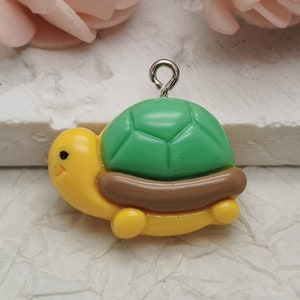 Puede incluir: Un pequeño amuleto de tortuga amarillo con caparazón verde y detalles marrones. La tortuga tiene un lazo plateado para sujetarlo a un collar o pulsera.