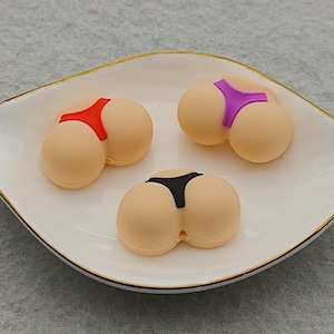 May include: Three novelty butt-shaped items with different coloured bikini bottoms, red, purple, and black, are displayed on a white, leaf-shaped dish with a gold trim. The items are beige in colour.