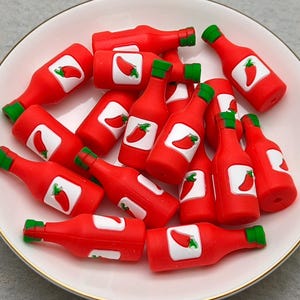 Puede incluir: Un plato blanco con borde dorado contiene numerosas botellas pequeñas de salsa picante roja. Cada botella tiene una tapa verde y una etiqueta blanca con una ilustración de un pimiento rojo. Las botellas están esparcidas por el plato.