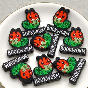 May include: A collection of colorful, cartoonish bookworm charms. Each charm features a red bookworm reading a book, with green leaves and the word "BOOKWORM" in white on a black background. The charms are arranged on a white plate with a gold rim.