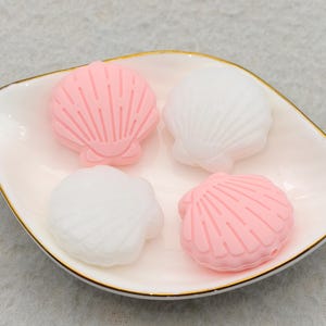 May include: A white ceramic dish with a gold rim holds four seashell-shaped objects. Two are pink, and two are white. The dish is leaf-shaped. The seashells have detailed ridges.