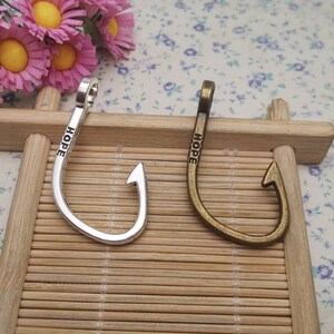 May include: Two metal fish hook charms, one silver and one bronze, each inscribed with the word "HOPE". The charms are displayed on a small wooden structure, with pink flowers in the background.