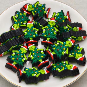 May include: A collection of Christmas-themed charms featuring a green character with a red and white Santa hat and a decorated Christmas tree. The charms are on a white plate with a gold rim.